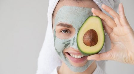 Close up of beautiful smiling girl cover face holding avocado looking at camera isolated on gray background. Happy pretty young woman advertising natural eco-friendly cosmetics for facial skincare.