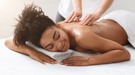 Therapist exfoliating black young woman body at spa, rubbing her back with scrub