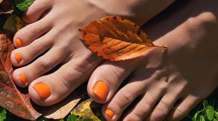Beautiful woman feet close up. Orange pedicure on the autumn background. Yellow and red fallen leaves.
